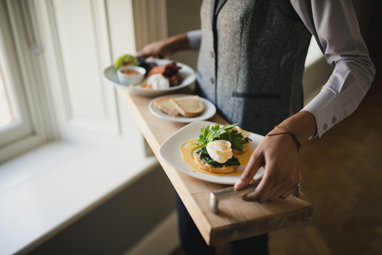A waiter is standing while holding a breakfast tray with a full English breakfast and poached eggs on toast covered in Hollandaise sauce on it.