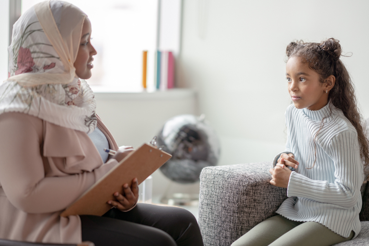 A Muslim Mother sits with her daughter as she teaches her from the comfort of their own home. The young girl is seated on a sofa while the mother sits across from her on a chair with a clipboard in her hands. She is testing the young girls memory and recording her answers.