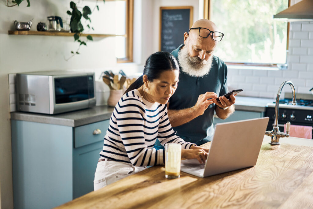 A mature couple using a laptop and phone in the kitchen at home. Husband and wife browsing online using social media apps with their wireless devices at home. Man and woman browsing, shopping online